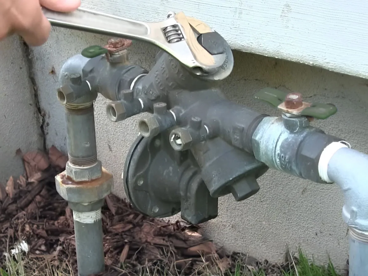 Wrench on a weathered backflow preventer at a house foundation, backflow prevention