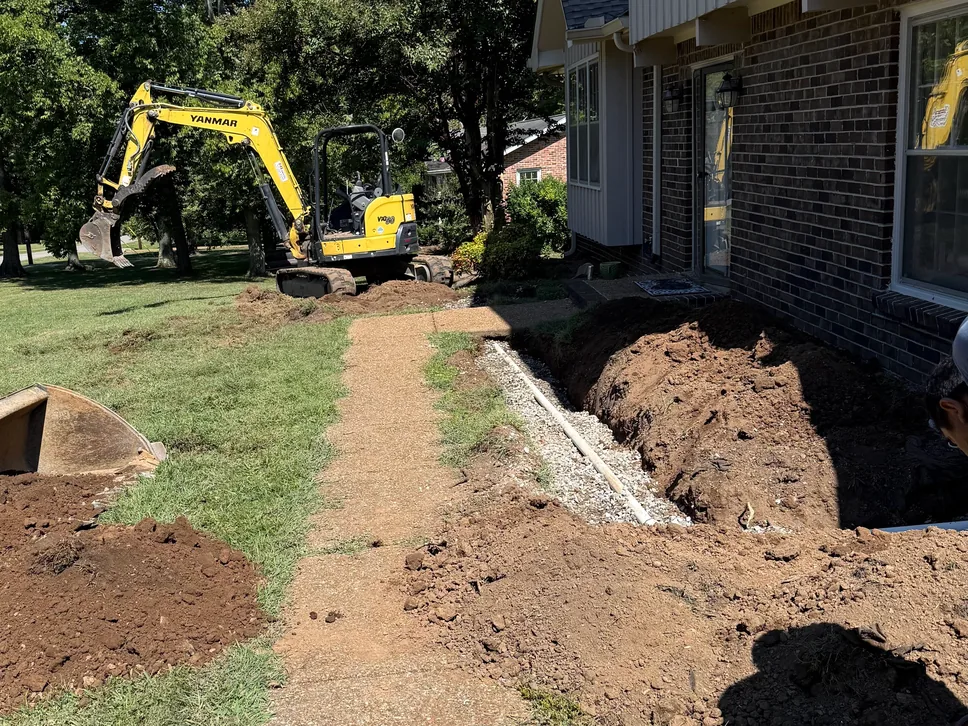 Mini excavator and a drain line trench along a brick house, sewer line repair